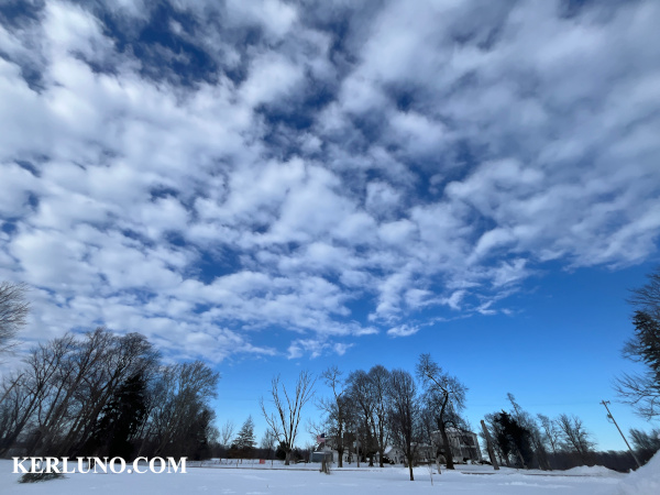 clear blue sky with some clouds, featuring snow on the ground with a house in the background.